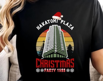 May include: Black t-shirt featuring a graphic of the Nakatomi Plaza building with a Santa hat, the words "Nakatomi Plaza Christmas Party 1988," and a sunset design. The bottom of the shirt reads "Christmas Hoodies & Sweatshirt Available."