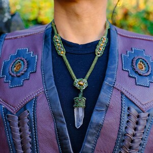 Könnte beinhalten: Eine grüne und braune Makramee-Kette mit einem Anhänger aus klarem Bergkristall. Die Kette wird von einer Person getragen, die eine lila und schwarze Lederweste trägt.