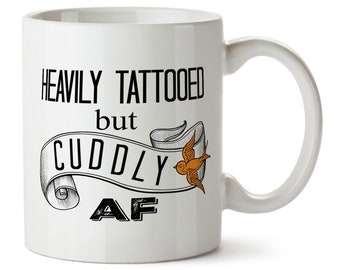 May include: White ceramic coffee mug with a black and white graphic that says "Heavily tattooed but cuddly AF" with a stylized bird illustration.