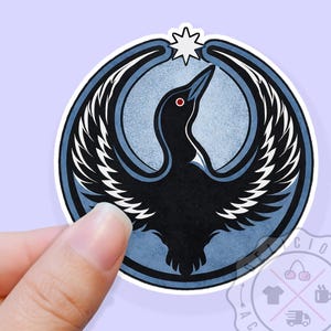 May include: A sticker featuring a stylized black bird with outstretched wings, set against a light blue background within a circular frame. A white star is above the bird's head. The sticker is held by a hand.