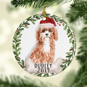 May include: A round ceramic Christmas ornament featuring a watercolor illustration of a brown and white dog wearing a red Santa hat. The dog is surrounded by a green wreath with red berries. The ornament also includes the text "DUDLEY 2019" on a white bone.