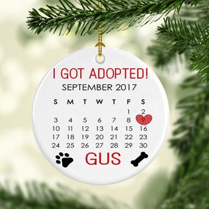 K&ouml;nnte beinhalten: Runder, wei&szlig;er Keramikschmuck mit dem Text "I GOT ADOPTED! SEPTEMBER 2017." Ein Kalender markiert den 2. September mit einem Herz. Enth&auml;lt einen Pfotenabdruck, einen Knochen und den Namen "GUS."