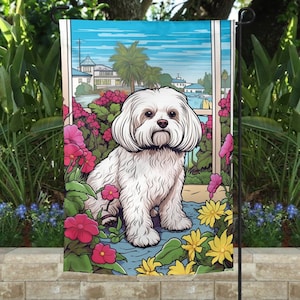 May include: A white Maltese dog sitting in a garden with pink and yellow flowers. The dog is looking at the camera. The background is a blue sky with a building in the distance.