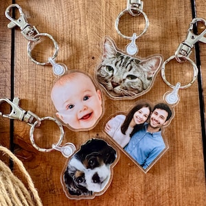 May include: A collection of personalized keychains featuring photos of a baby, a cat, a couple, and a dog. Each keychain has a silver-colored clasp and a split ring. The keychains are arranged on a wooden surface.