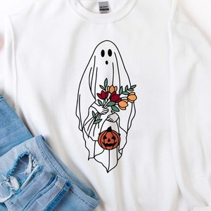 K&ouml;nnte beinhalten: Wei&szlig;es Sweatshirt mit einer Geisterillustration, die Blumen und einen K&uuml;rbis h&auml;lt. Das Geisterdesign ist schwarz umrandet. Das Bild enth&auml;lt auch eine Sonnenbrille und eine blaue Jeans.