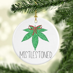 May include: White ceramic Christmas ornament with a green glitter cannabis leaf design and a red bow. The text "MISTLESTONED" is printed in black. The ornament hangs from a gold string.