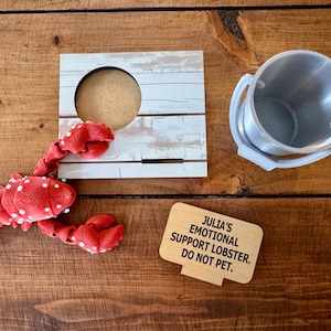 May include: A red lobster figurine with white spots, a wooden picture frame, a small metal bucket, and a wooden sign that reads "Julia's Emotional Support Lobster. Do Not Pet." on a wooden surface.