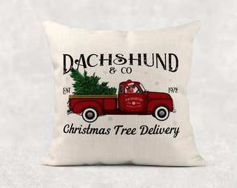 May include: Cream-colored throw pillow featuring a red vintage truck with a Christmas tree in the bed. The truck has a dachshund wearing a Santa hat in the driver's seat. Text reads "DACHSHUND & CO" and "Christmas Tree Delivery."