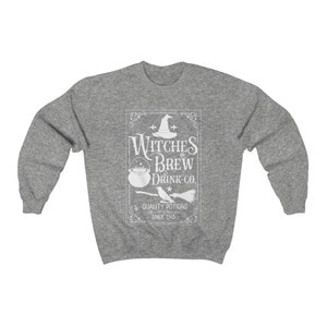 May include: A heather gray sweatshirt featuring a white graphic that reads "Witches Brew Drink Co." with a witch's hat, cauldron, and broom. The text "Quality Potions Since 1543" is also included in the design. The sweatshirt has long sleeves and a crew neck.