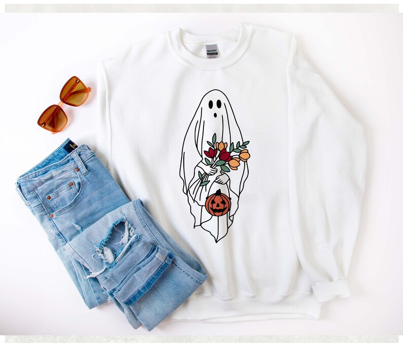 K&ouml;nnte beinhalten: Wei&szlig;es Sweatshirt mit einer Geisterillustration, die Blumen und einen K&uuml;rbis h&auml;lt. Das Geisterdesign ist schwarz umrandet. Das Bild enth&auml;lt auch eine Sonnenbrille und eine blaue Jeans.