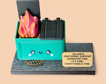 May include: A whimsical sculpture of a green dumpster with a cartoon face, a black lid, and a flame inside. The sculpture sits on a gray base with a sewer grate detail and a small plaque that reads "Jillian's Emotional Support Dumpster Fire. It's Fine. Everything's Fine."