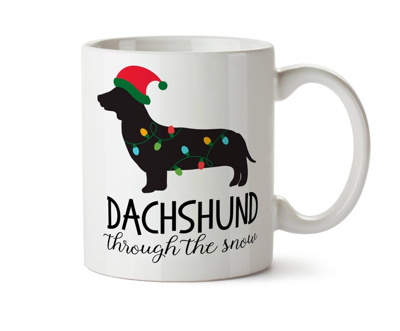 Puede incluir: Taza de cer&aacute;mica blanca con la silueta de un perro salchicha negro con un gorro de Pap&aacute; Noel y luces navide&ntilde;as. La taza tiene el texto "DACHSHUND through the snow" en escritura negra. La taza tiene un asa blanca.