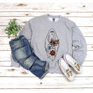 K&ouml;nnte beinhalten: Hellgraues Sweatshirt mit einem schwarz-wei&szlig;en Geisterdesign, das Blumen und einen K&uuml;rbis h&auml;lt. Das Sweatshirt wird mit blauen Jeans und wei&szlig;en Turnschuhen kombiniert, vor einem wei&szlig;en Holzhintergrund.
