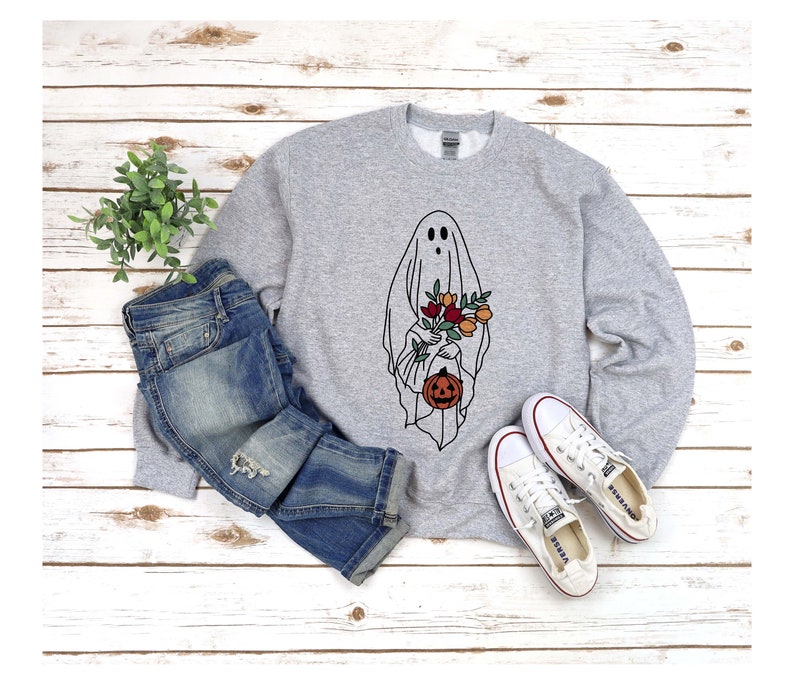 K&ouml;nnte beinhalten: Hellgraues Sweatshirt mit einem schwarz-wei&szlig;en Geisterdesign, das Blumen und einen K&uuml;rbis h&auml;lt. Das Sweatshirt wird mit blauen Jeans und wei&szlig;en Turnschuhen kombiniert, vor einem wei&szlig;en Holzhintergrund.