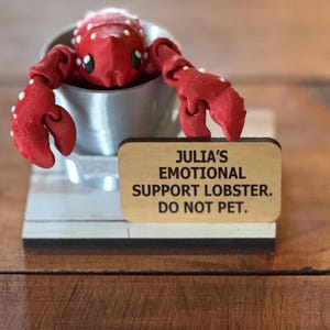 May include: A red lobster figurine with white accents sits in a silver container. The lobster is on a wooden base with a sign that reads "JULIA'S EMOTIONAL SUPPORT LOBSTER. DO NOT PET."