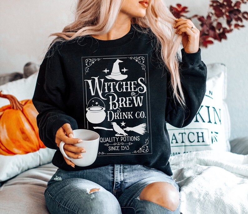 May include: Black sweatshirt with a white graphic that reads "Witches Brew Drink Co." featuring a witch's hat, cauldron, and broom. The text "Quality Potions Since 1543" is also included in the design. The sweatshirt is a Halloween-themed apparel item.