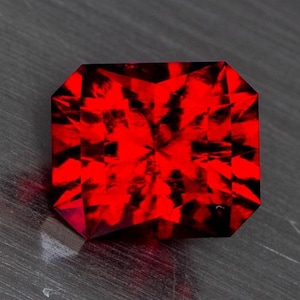 May include: A deep red gemstone with a square cut and a faceted design. The gemstone is set against a gray background.
