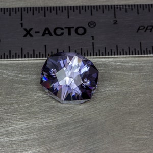 May include: A purple gemstone with a faceted cut, measuring approximately 1 inch in diameter, is displayed on a silver surface next to a ruler labeled "X-ACTO" and "PRECISION".