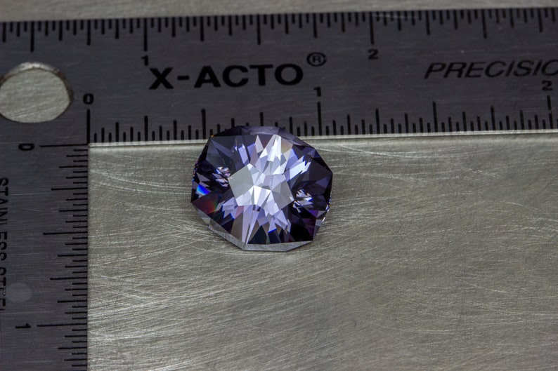 May include: A purple gemstone with a faceted cut, measuring approximately 1 inch in diameter, is displayed on a silver surface next to a ruler labeled "X-ACTO" and "PRECISION".