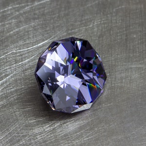 May include: A large, faceted, purple gemstone with a slightly iridescent sheen. The gemstone is on a brushed metal surface.