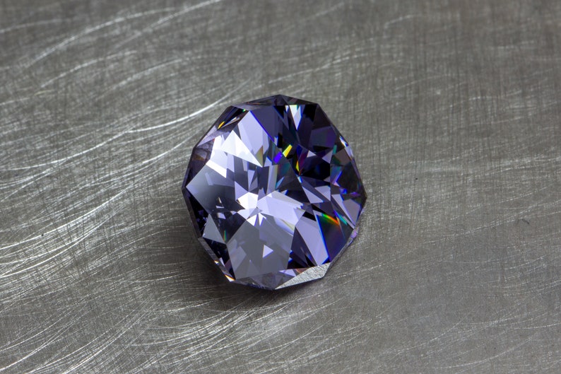 May include: A large, faceted, purple gemstone with a slightly iridescent sheen. The gemstone is on a brushed metal surface.