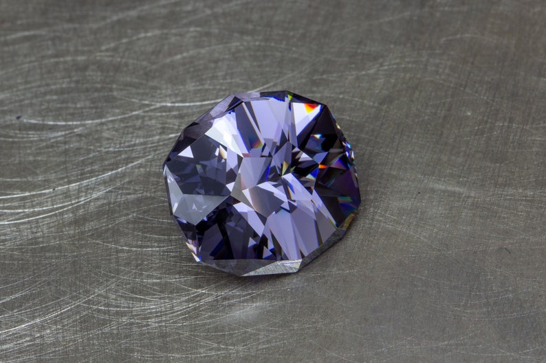 May include: A large, faceted, purple gemstone with a dark, almost black, hue. The gem is cut in a unique, octagonal shape and has a high degree of brilliance.