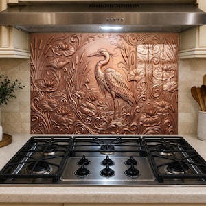 May include: A copper-toned metal backsplash panel featuring a relief design of a heron standing among water lilies and cattails. The panel is installed behind a stainless steel gas cooktop with five black burners and a matching range hood.