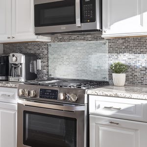 May include: A stainless steel gas range with a matching over-the-range microwave, positioned below white cabinets. The kitchen features a gray and white mosaic tile backsplash, a granite countertop, and a small potted green plant on the side.