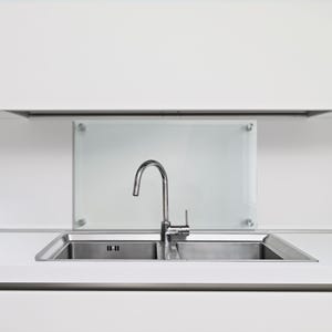 May include: A stainless steel double basin kitchen sink with a high-arc chrome faucet. Behind the sink is a rectangular frosted glass backsplash panel mounted with four circular silver hardware fasteners against a plain white wall.