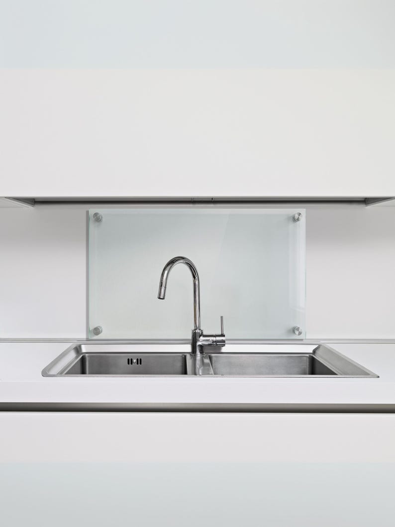 May include: A stainless steel double basin kitchen sink with a high-arc chrome faucet. Behind the sink is a rectangular frosted glass backsplash panel mounted with four circular silver hardware fasteners against a plain white wall.