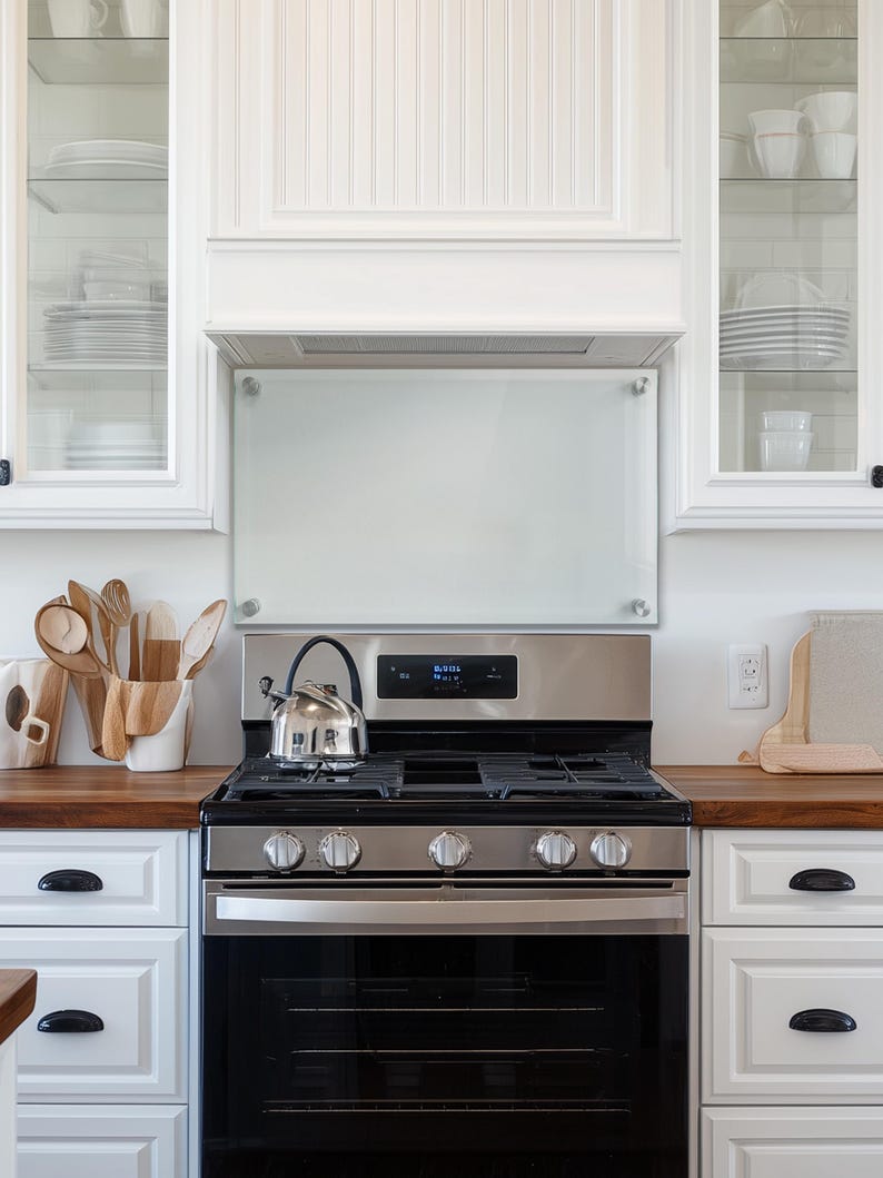 May include: A rectangular glass kitchen backsplash panel mounted with four silver hardware standoffs, positioned above a stainless steel gas range with a black oven door and a silver tea kettle resting on the stovetop burner grates.