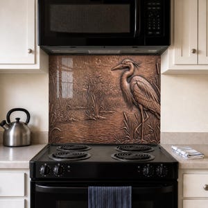 May include: A copper-toned metal backsplash panel featuring a relief sculpture of a heron standing in a marshy landscape with reeds and cattails. The rectangular panel is installed behind a black electric stove and below a black microwave.
