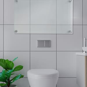 May include: A rectangular clear glass dry-erase board mounted with four silver-toned metal standoffs on a tiled wall. Below the board is a rectangular silver flush plate centered on the wall, positioned above a white wall-mounted toilet.