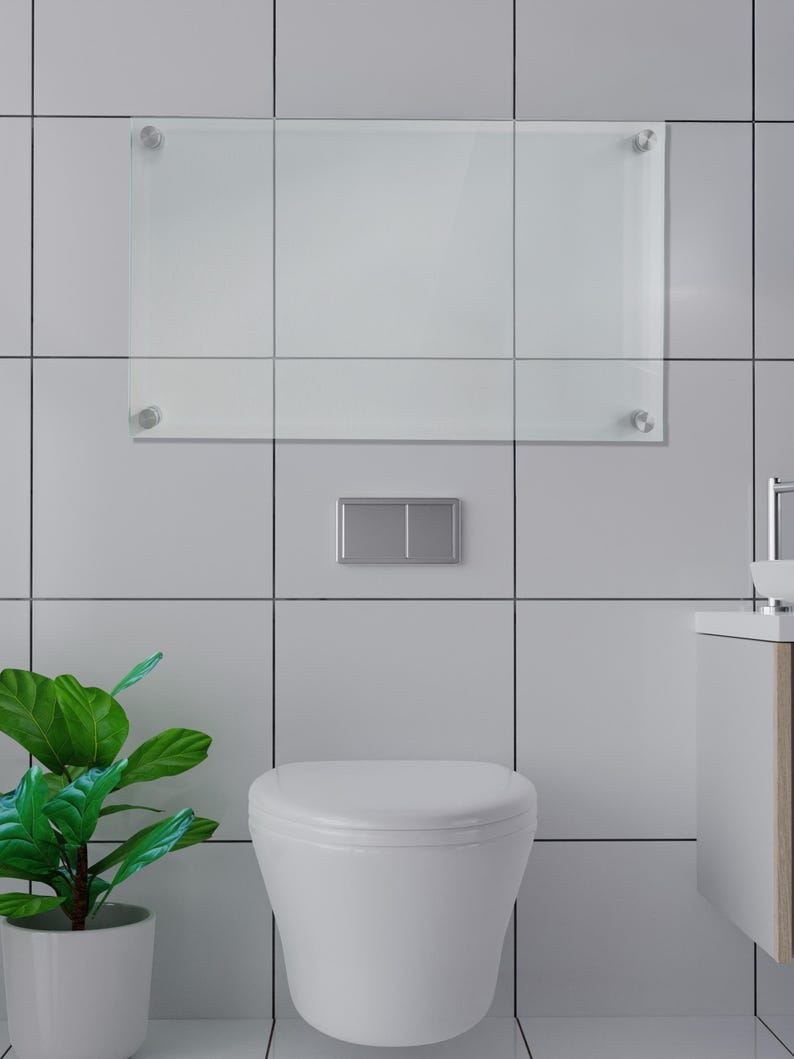 May include: A rectangular clear glass dry-erase board mounted with four silver-toned metal standoffs on a tiled wall. Below the board is a rectangular silver flush plate centered on the wall, positioned above a white wall-mounted toilet.