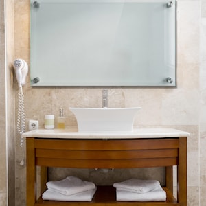 May include: A rectangular wall-mounted mirror with frosted glass and four silver-toned metal standoffs at the corners, positioned above a white vessel sink, a wooden vanity cabinet with two drawers, and two shelves holding folded white towels.