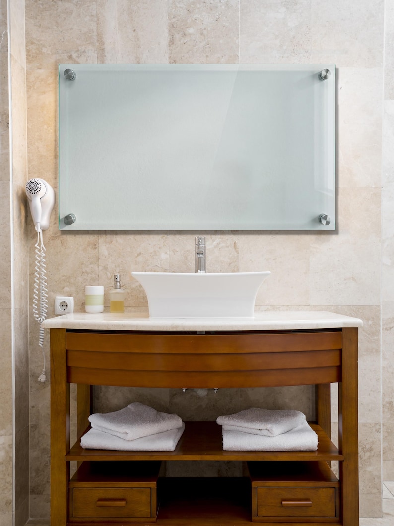May include: A rectangular wall-mounted mirror with frosted glass and four silver-toned metal standoffs at the corners, positioned above a white vessel sink, a wooden vanity cabinet with two drawers, and two shelves holding folded white towels.