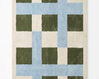 May include: A rectangular rug with a geometric pattern. The rug features a cream-colored background with a grid design of olive green squares and light blue intersecting lines. The rug is made of a soft, plush material.