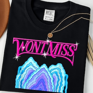 Può includere: T-shirt nero girocollo con la scritta rosa "WONT MISS" con stelle bianche e una grafica a strati blu, viola e bianca. Una collana d'oro con pendente è appoggiata sulla maglietta. Un capo d'abbigliamento casual.