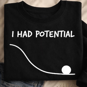 Pode incluir: Uma caneca de cerâmica branca com uma alça preta e o texto "But First, Coffee" em negrito preto. A caneca está sobre um fundo desfocado, sugerindo um ambiente acolhedor.