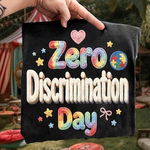 May include: Black fabric with the words "Zero Discrimination Day" in a colorful, textured design. The word "Zero" is in rainbow colors, and the design includes stars, a heart, and a puzzle piece.