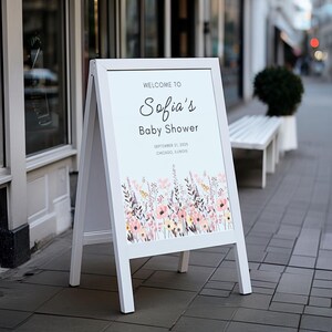 May include: A white A-frame sign with the text "Welcome to Sofia's Baby Shower" in elegant script. The sign features a floral design at the bottom and is displayed outdoors on a city sidewalk.
