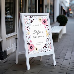 May include: A white A-frame sign with a floral design and the text "Welcome to Sofia's Baby Shower." The sign features pink and yellow flowers with green and brown foliage. The date and location are also displayed.