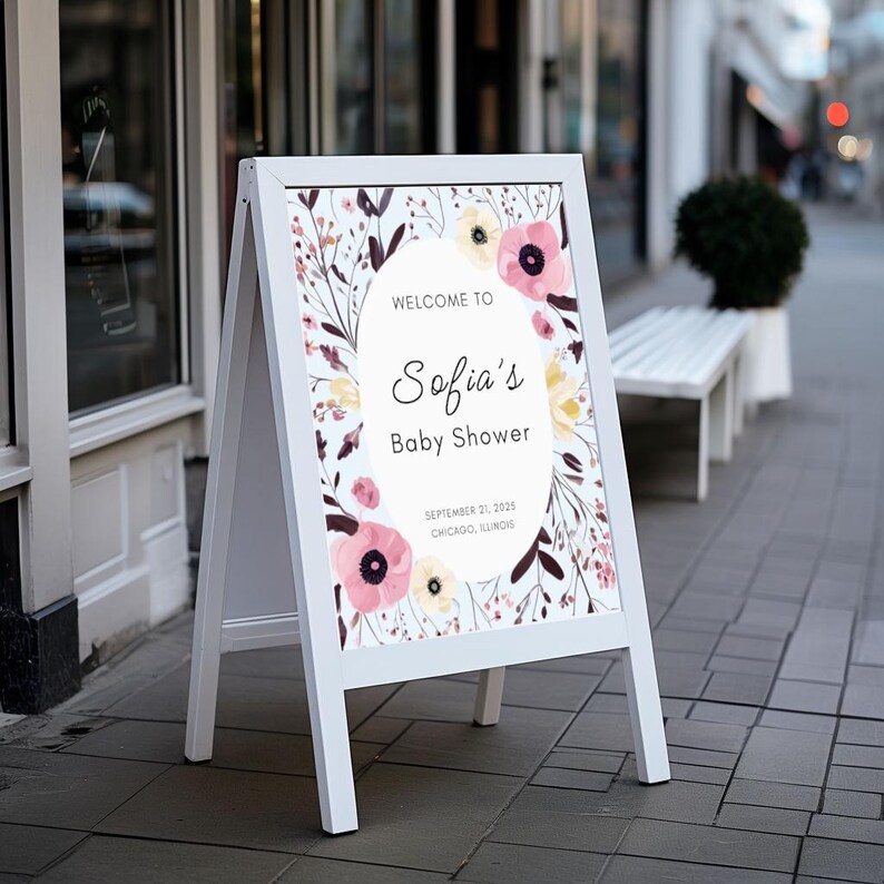 May include: A white A-frame sign with a floral design and the text "Welcome to Sofia's Baby Shower." The sign features pink and yellow flowers with green and brown foliage. The date and location are also displayed.