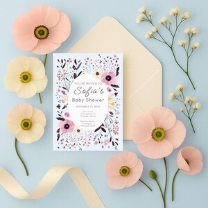 May include: A baby shower invitation with floral design, surrounded by peach and cream-colored paper flowers and a cream envelope. The invitation reads "Sofia's Baby Shower" with event details. A cream ribbon is at the bottom.