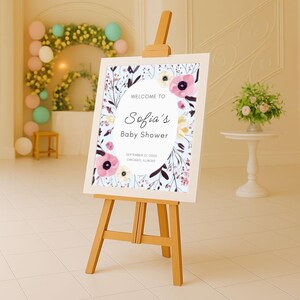 May include: A wooden easel displays a baby shower sign. The sign features floral illustrations and the text "Welcome to Sofia's Baby Shower" with the date and location. The sign is framed with a light blue background and floral accents.