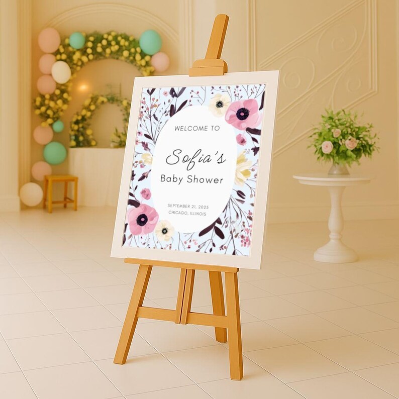 May include: A wooden easel displays a baby shower sign. The sign features floral illustrations and the text "Welcome to Sofia's Baby Shower" with the date and location. The sign is framed with a light blue background and floral accents.