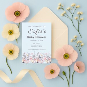 May include: A baby shower invitation with the text "Sofia's Baby Shower" on a light blue background, surrounded by pink and yellow paper flowers, a cream envelope, and sprigs of white flowers. A cream ribbon is also present.