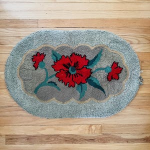 May include: An oval-shaped tufted rug featuring a central gray scalloped panel decorated with a large red flower and green leaves. The outer border of the rug is a light gray color, and the entire piece has a textured, looped pile construction.