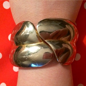 May include: A gold-colored cuff bracelet with a rounded, abstract design. The bracelet is worn on a wrist, reflecting the surrounding environment. The background is a red fabric with white polka dots.