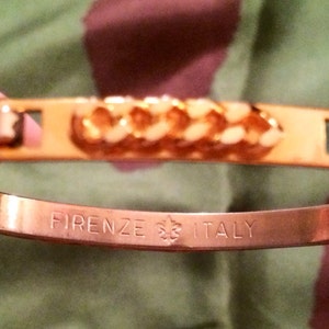 May include: Gold-toned bangle bracelet with a central chain design and brown leather accents. The bracelet is engraved with "FIRENZE ITALY".